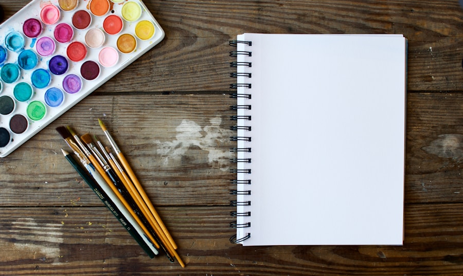 A notebook, paints and brushes on a wooden table.