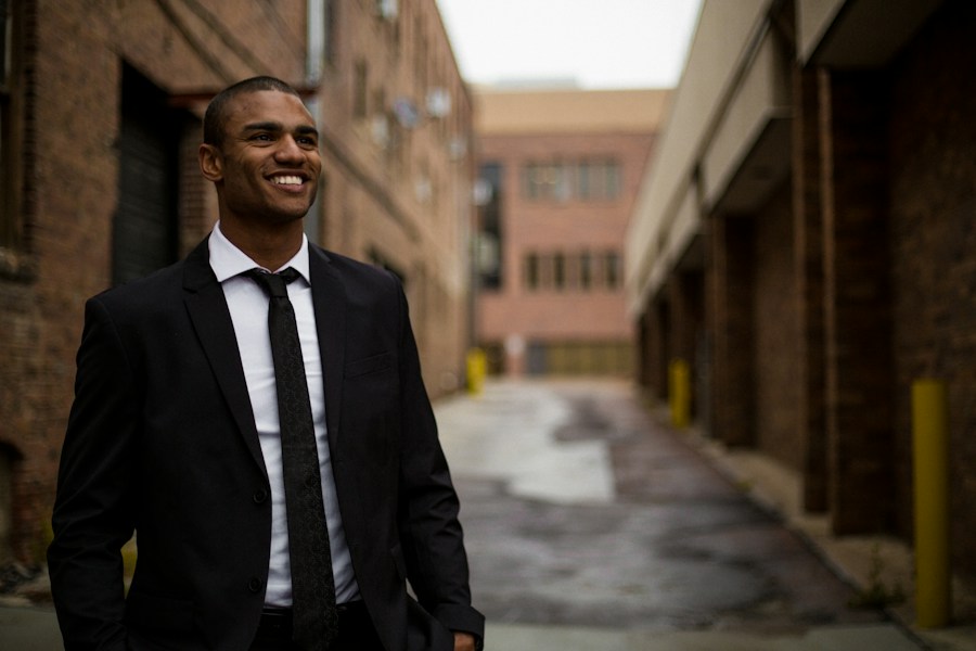 A young man in a suit smiles in an alleyway.