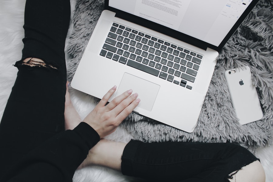 Person using a laptop with a smartphone beside them, sitting on a fur rug with ripped jeans.