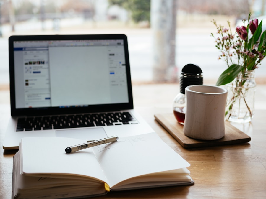 A laptop open on a social media page, a notebook with a pen, and a cup of tea on a wood table.