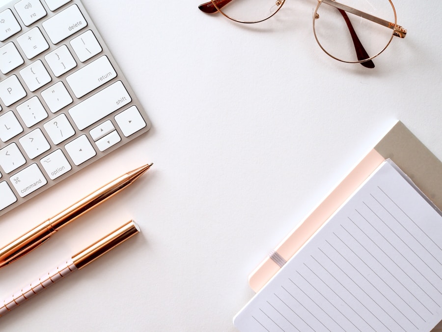 A minimalist workspace with a keyboard, glasses, two pens, and a notepad on a white desk.