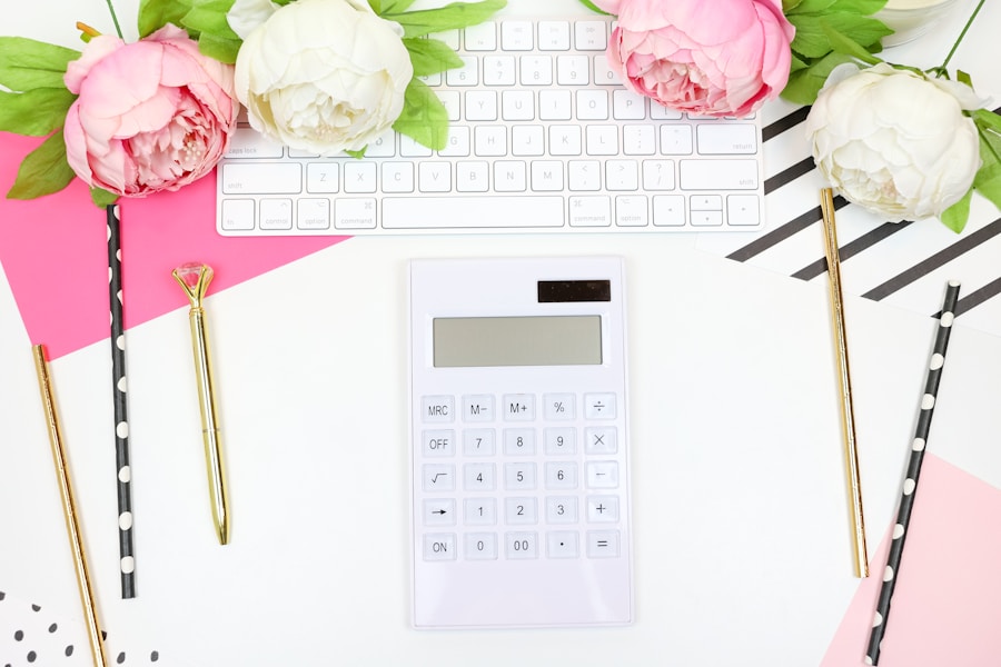 A desk with a white calculator, keyboard, and decorative items including peonies and stylish stationery on a pink and white background.