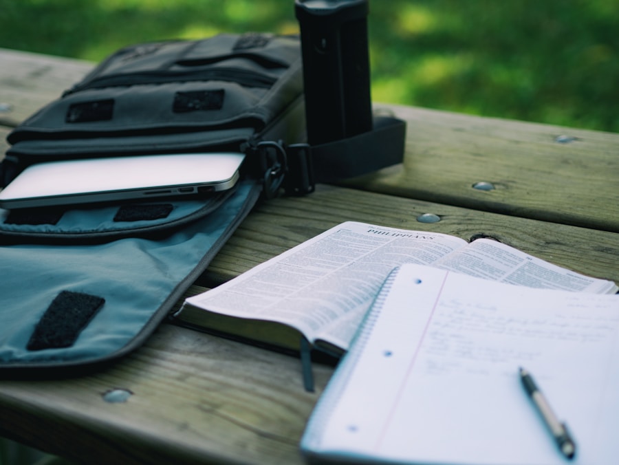 A study setup outdoors with an open book, notebook, pen, and a laptop in a bag on a wooden table.