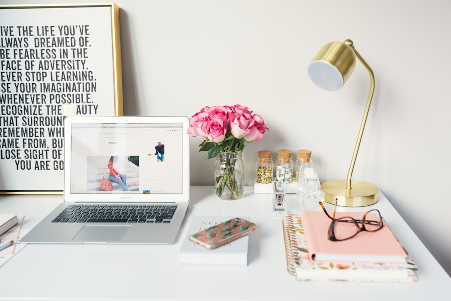 A well-organized desk with a laptop, notebook, glasses, a vase of pink roses, and a decorative lamp.