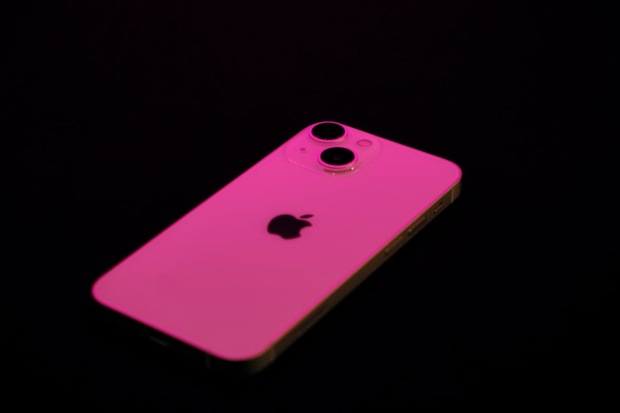 A smartphone with a pink hue rests on a black surface, displaying the Apple logo and dual rear cameras.