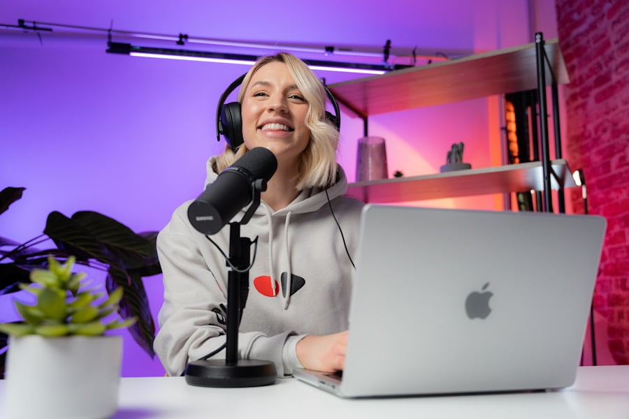 Person wearing headphones sits at a desk with a laptop and microphone, smiling. The background has colorful lighting and a shelf with various items.