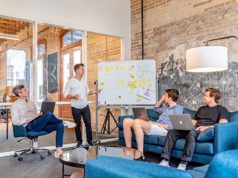 Four people in a modern office are engaged in a meeting. One person is standing by a whiteboard with sticky notes, while the other three are seated on couches with laptops, attentively listening.