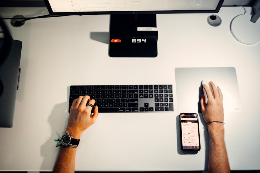 Person seated at a white desk using a desktop computer, operating a smartphone placed beside the keyboard, with both hands visible. A monitor, mouse, and several objects are also present on the desk.