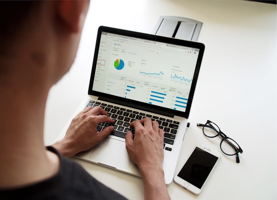 Person analyzing data on a laptop, with graphs and charts displayed on the screen. Nearby, there are eyeglasses and a smartphone on the desk.