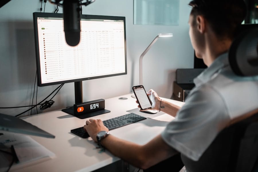 A person sitting at a desk is working on a computer and looking at a smartphone. The desktop has a monitor, keyboard, mouse, desk lamp, microphone, and digital clock displaying 6:34.