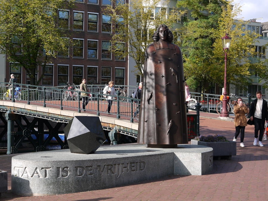 Bronze statue of a person next to a geometric sculpture on a stone pedestal, with a bridge and buildings in the background. Text on the pedestal reads, "STAAT IS DE VRIJHEID.