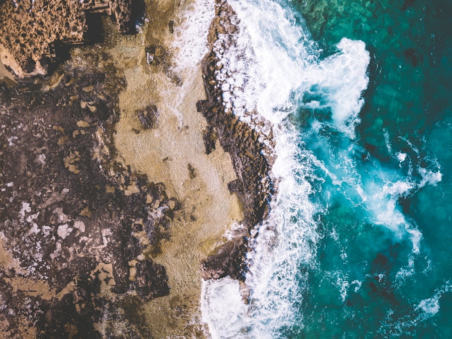 Aerial view of a rocky shoreline with waves crashing onto the shore and turquoise water blending with darker ocean sections.