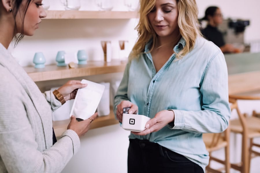 Two women in a store making a transaction with a square card reader. One woman is holding a receipt, and the other is holding a small device. Shelves with cups are in the background.