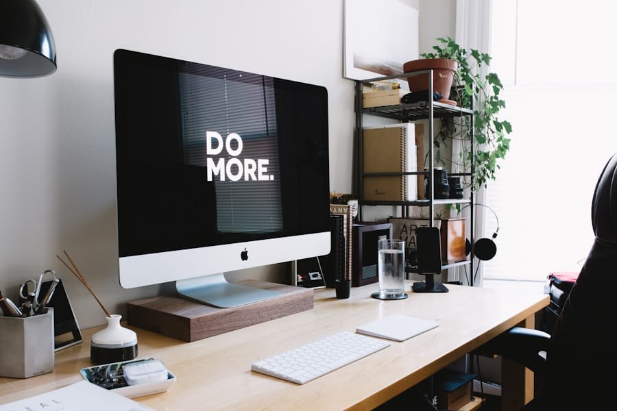 A desktop computer on a wooden desk displays the text "DO MORE." The workspace includes books, a water glass, and a potted plant.